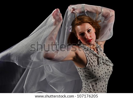 A beautiful young woman arches her back as she holds the sheer fabric up over her head and the wind begins to take it away while isolated on a black background.