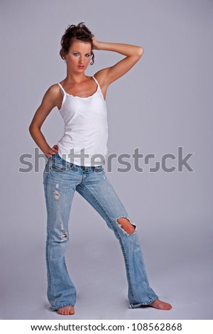 Beautiful young woman in torn blue jeans on a gray background with one hand on her hip and other hand holding her head.