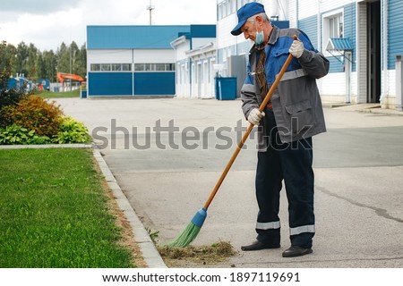 This is an elderly janitor with a broom in a medical mask on the street sweeping the territory. An old man in a work uniform works during a time of pandemic and unemployment. Foto stock © 
