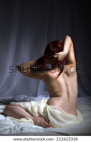 Back view of a naked woman sitting in bed playing with her hair