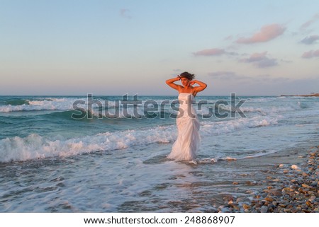 Barefoot young bride in a partially wet  wedding dress, enjoys walking in the water on a sandy beach in late summer, at dusk.