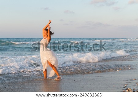 Barefoot young woman in a long white partially wet dress, enjoys a lonesome walk in the water, on a sandy beach in a late summer day, at dusk.