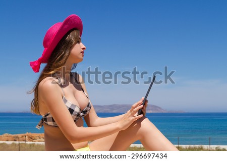 Gorgeous young busty naughty long haired brunette teen girl in swimwear enjoying the sun on a fresh spring morning by the ocean, poses for selfies using her tablet.
