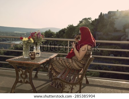 Woman with Red Head Cloth is Drinking Coffee on Balcony with River Background