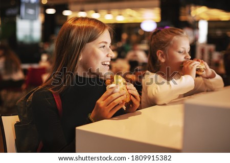 Foto Bild Hübsches Schulmädchen isst Hamburger und sitzt drinnen in einem Cafe. Glückliches Kind, das Junkfood in einem Restaurant isst. Seitenansicht