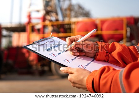 Action of safety office is writing on checklist paper during safety audit and risk verification at drilling site operation with blurred background of mount truck rig. Selective focus at hand.