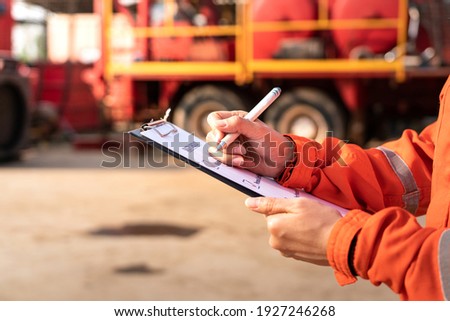 Action of safety officer is check on checklist document during safety audit and risk verification at drilling site operation with blurred background of mount truck rig. Selective focus at hand.