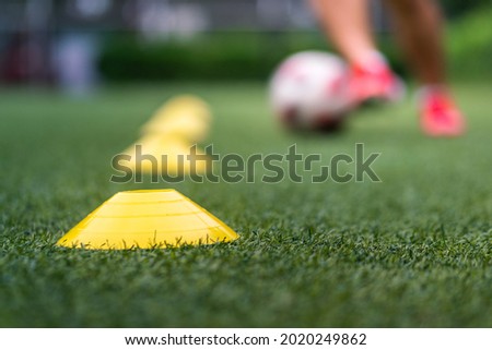 A yellow obstacle cone (Focus), sport training equipment on artificial pitch ground with action of a football player is practice to dribbling the ball as blurred background. Sport training concept. 商業照片 © 