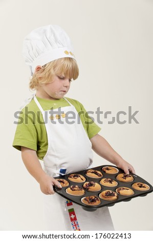 A young boy with a chef hat on is holding a tray of muffins.