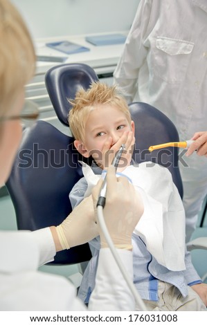 A boy in a dental chair covers his mouth as the dentist approaches with a tool.