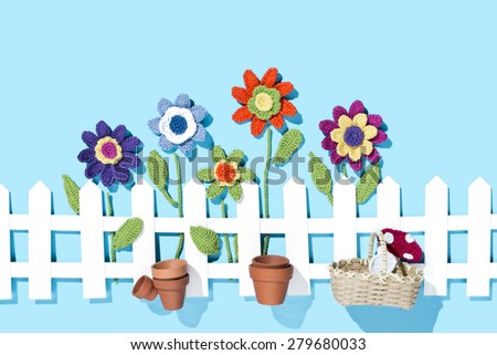 crochet flowers behind a white paper fence with flower pots, basket and a mushroom on blue background