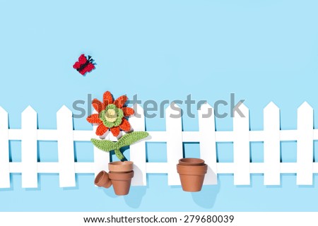 crochet flower with pots, a butterfly and a white fence on blue background