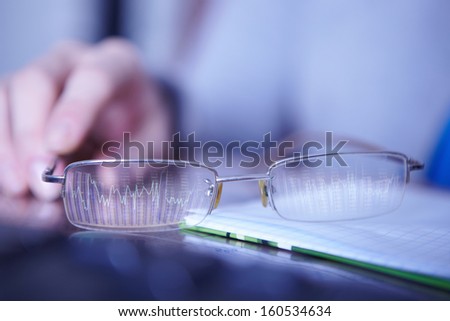 Hand of financial advisor with glasses lying on the desktop before the monitor with charts.