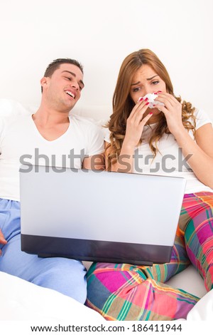 ordinary casual young couple in love watching sad tragic and dramatic movie in bed on laptop computer. Man laughing at woman with tissue while she is crying and shedding tears