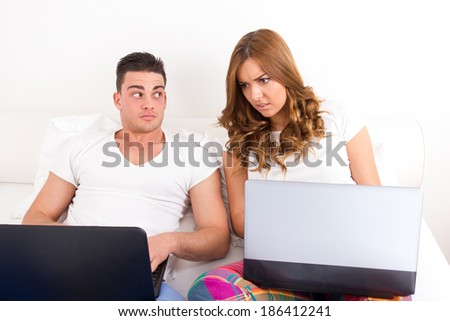 depressed and shocked young woman looking at man\'s computer. Partners in bed with laptop computers. Caught in cheating on a social network
