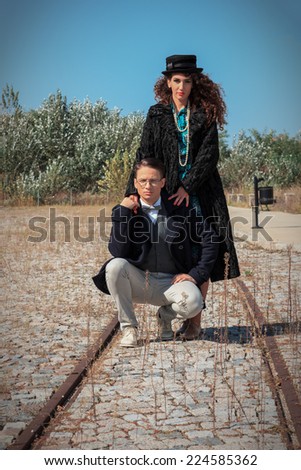 Retro fashionable woman and man standing on railroad in vintage style of old town, Girl wearing coat and hat for cold windy weather standing behind her man