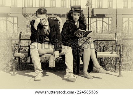 Young fashionable couple in quarrel sitting on bench in old town reading book in vintage style