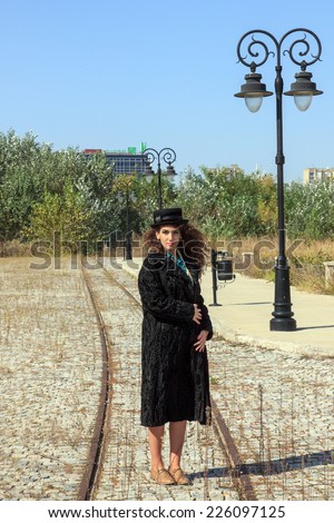 Woman with curly hair wearing retro fur coat and hat standing on railway in vintage town