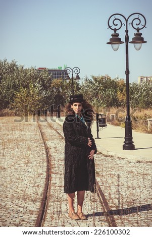 Brunette retro woman wearing coat and hat standing on railroad by lamppost