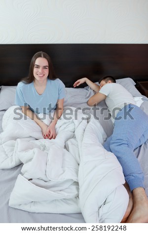 Young cute caucasian couple, man and woman in bed, female smiling and looking at camera while male sleeping. Domestic morning atmosphere