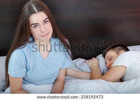 Dissatisfied beautiful young woman in bed with sad expression on her face, sex problems in long relationship or marriage while man sleeping. Saturation or satiety between caucasian couple, disinterest