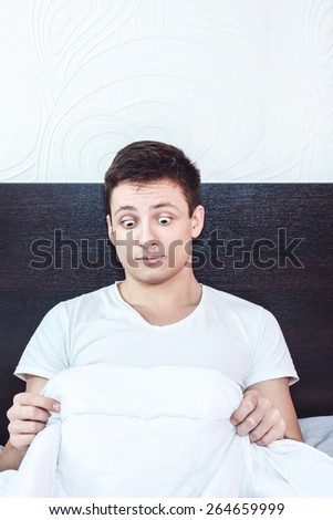 surprised and shocked good looking young man in bed looking down at his penis under white covers sheet in bedroom. Concept photo of male sexuality and man sex peny problems, domestic atmosphere.