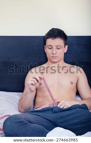 Young handsome half-naked man holding tape measure and measure desired penis length or size while sitting on bed in bedroom in pajamas. Male model showing suspicious length of his pride.