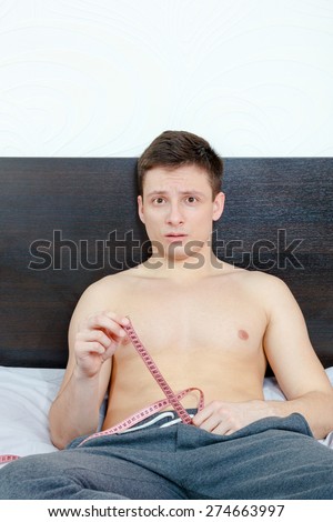 Young handsome half-naked man holding tape measure and measure desired penis length or size while sitting on bed in bedroom in pajamas. Frightened and insecure male model showing length of his pride.