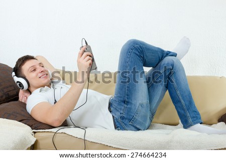 Young man smiling enjoying music with headset and cellphone , Guy relaxing on couch at home listening to music over mobile phone with earphones