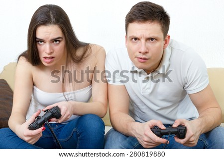 Young couple sitting in living room and play video games on console or pc with joysticks while looking in screen or TV. Lifestyle photo of man and woman in jeans at home having fun or entertainment.