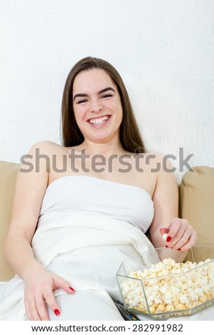 Girl watching movie or TV laughing having fun eating popcorn in bed. Alone young Caucasian woman model sitting down on bed.  Smiling female watching funny comedy in living room at home.
