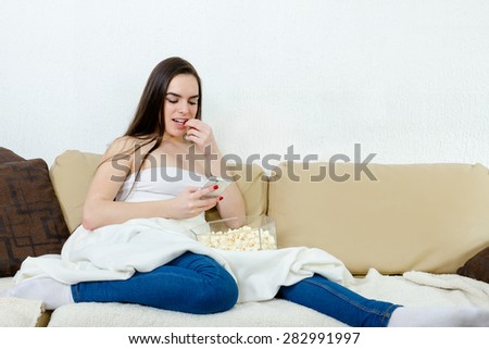 Young beautiful women typing message on mobile phone and eating popcorn. Alone happy female model with cellphonel sitting on bed with blanket.  Smiling girl watching screen in living room at home.