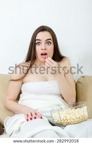 Woman scared funny watching scary movie eating popcorn. Beautiful girl watching TV in bed on white domestic background. Shocked young female watching horor while sitting on sofa at home.