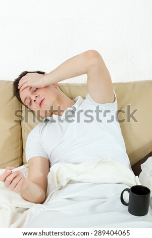 Portrait of sick man in bedroom suffering from influenza measuring temperature with thermometer and drinking tea