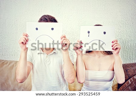Sad young couple. Unhappy young couple  in despair sitting on couch. Male and female with sad faces. Man and woman cover their faces with sad smile drawn on paper with one tear.