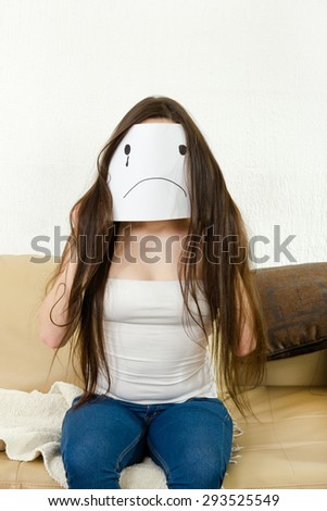 Adult girl cover her face with sad smile drawn on paper with one tear. Woman and love problems. Sad teenager in despair. Unhappy young person  sitting on couch. Female with disappointed smiley face.