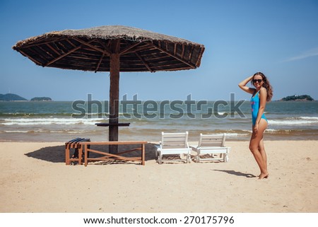 Happy woman resting on a tropical beach near deck chair and sun umbrella. Blue sky, deserted beach