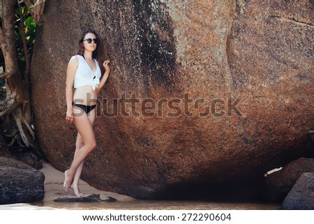 Young girl in black swimsuit and white blouse on the rocks near the sea beach.full-length portrait