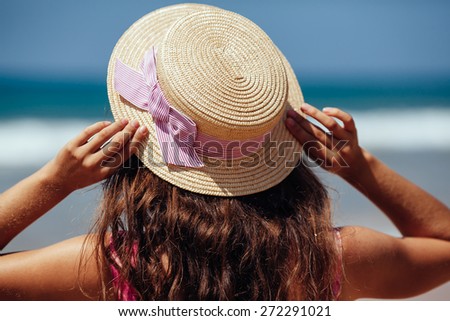 Young woman standing on a beach holding her straw hat and looking to the horizon.