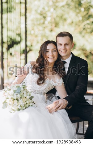 Stylish groom in a gray checkered suit and a cute blonde bride in a white lace dress with a long veil are hugging in the park near the old house. Wedding portrait of smiling newlyweds. ストックフォト © 