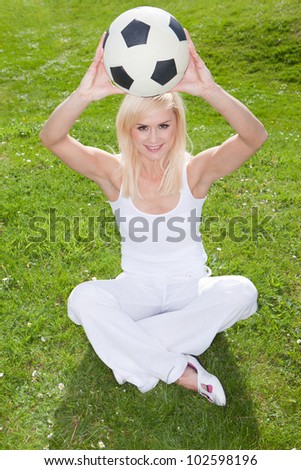 Smiling blonde woman sitting on green grass holding a soccerball or football above her head
