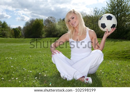 Smiling blonde woman sitting on green grass in the park about to throw a football which she is holding in her left hand