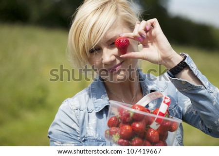 Attractive blonde woman holding up large a ripe red strawberry in her fingers from a punnet full that she is holding outdoors in sunshine