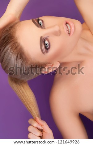 Head and shoulders portrait of a naked sexy woman with beautiful eyes holding her blonde ponytail in her hand on a purple studio background