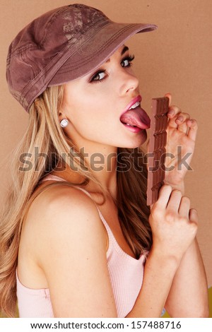 Sensual beautiful young woman wearing a brown peaked cap licking a chocolate bar while glancing across at the camera