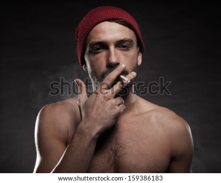 Atmospheric head and shoulders portrait of a shirtless man wearing a red beanie hat smoking a cigarette against a dark background