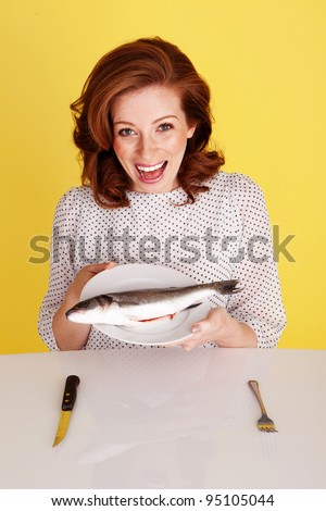 Humorous photo of a laughing redhead woman seated at a table holding a whole raw fish on her plate.