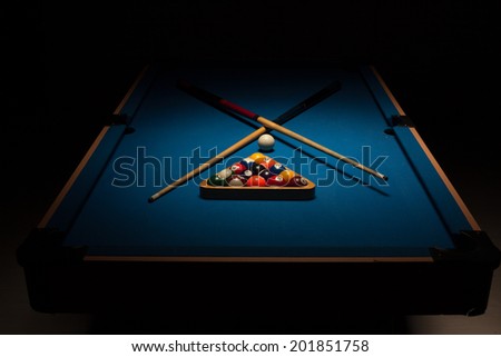 Pool equipment ready for a game with crossed wooden cues, racked balls and a cue ball on a blue baize table surrounded by darkness