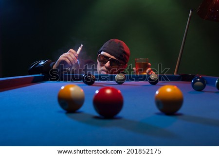 Man in sunglasses and a cap playing pool in a shadowy nightclub standing surveying the lay of the balls on the table smoking e-cigarette or e-shisha