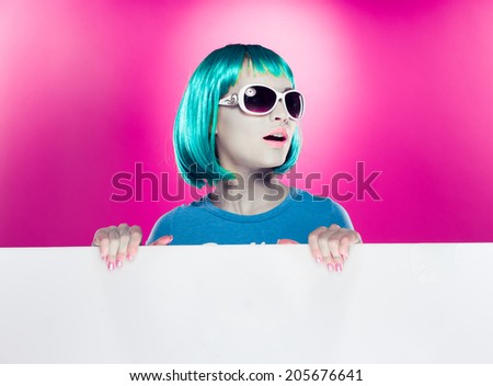 Trendy beautiful young modern woman with green hair wearing stylish sunglasses holding a blank sign with copyspace for your text over a magenta background, looking away